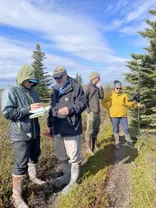 Four people stand near each other. One of them is holding a Trimble instrument and another is looking in their field notebook. They are standing on mixed foliage and there are a few evergreen trees nearby. The sky is partially cloudy.