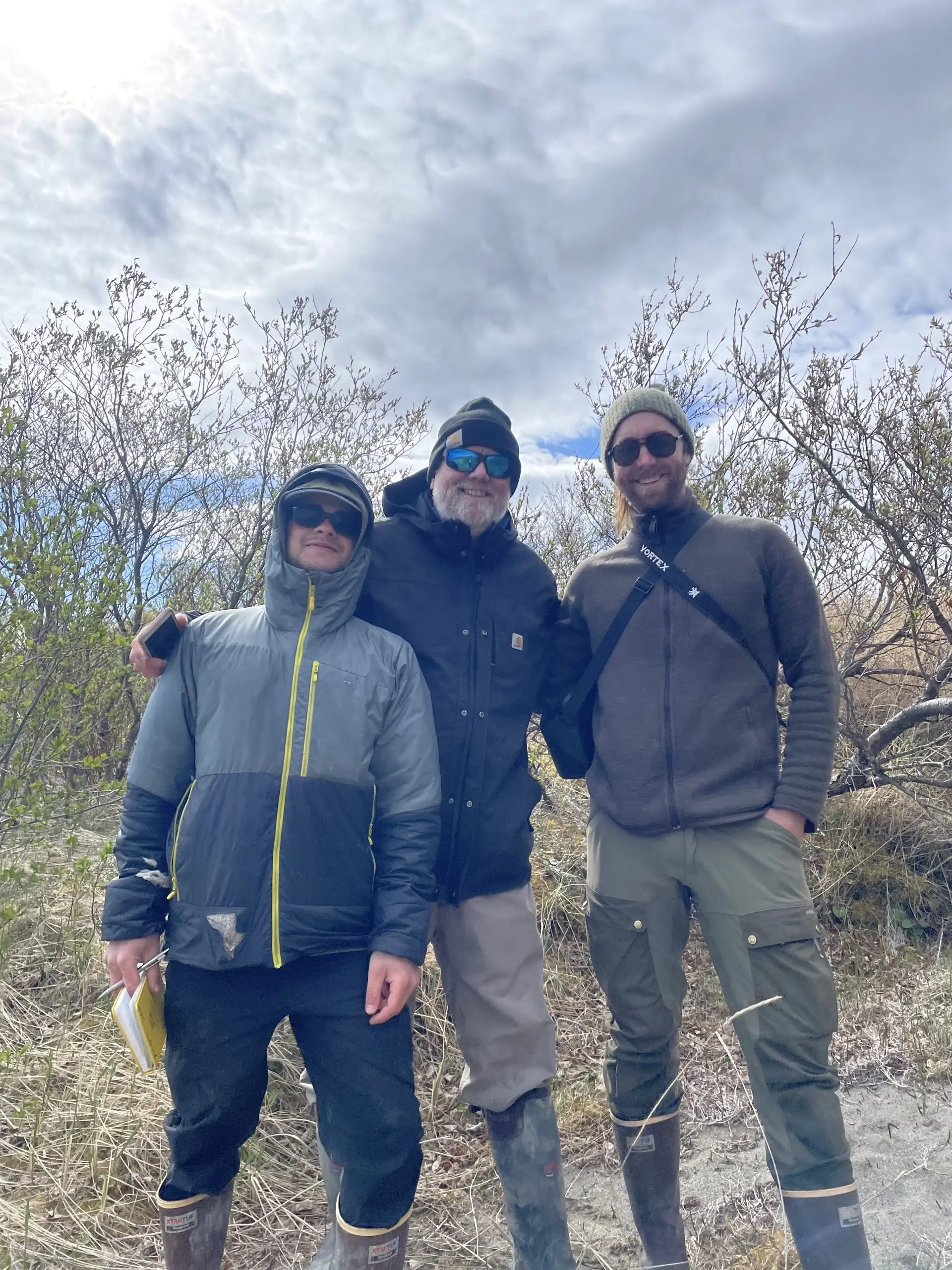 Three people stand near each other for a small group photo. They are standing in a sandy patch near some brush. The sky is cloudy.