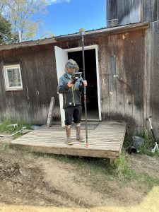 Seth Classen stands in front of a weathered wooden shack. He is holding a Trimble instrument. He is standing outside on a wooden porch, wearing a mosquito mask. 