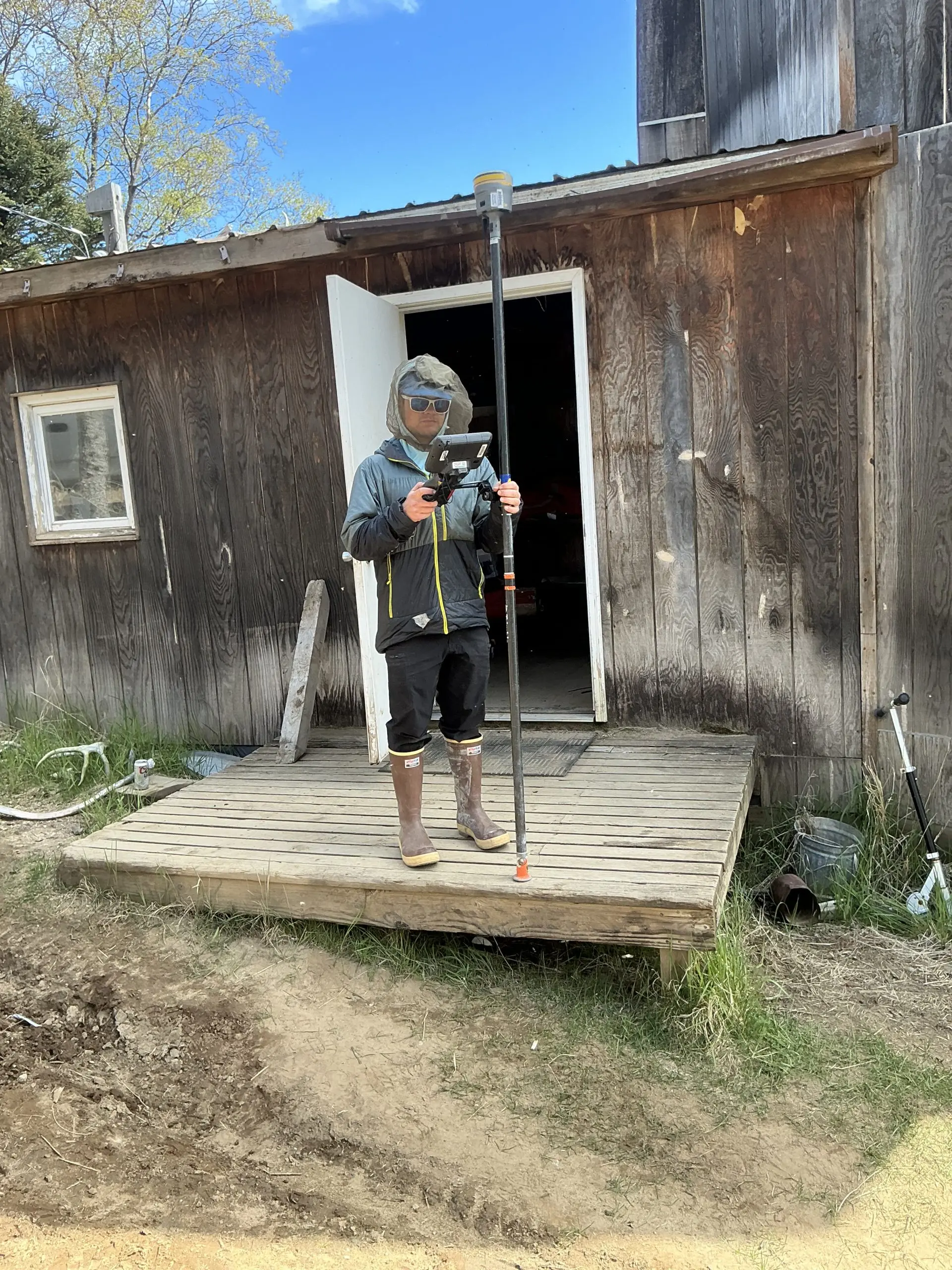 Seth Classen stands in front of a weathered wooden shack. He is holding a Trimble instrument. He is standing outside on a wooden porch, wearing a mosquito mask. 