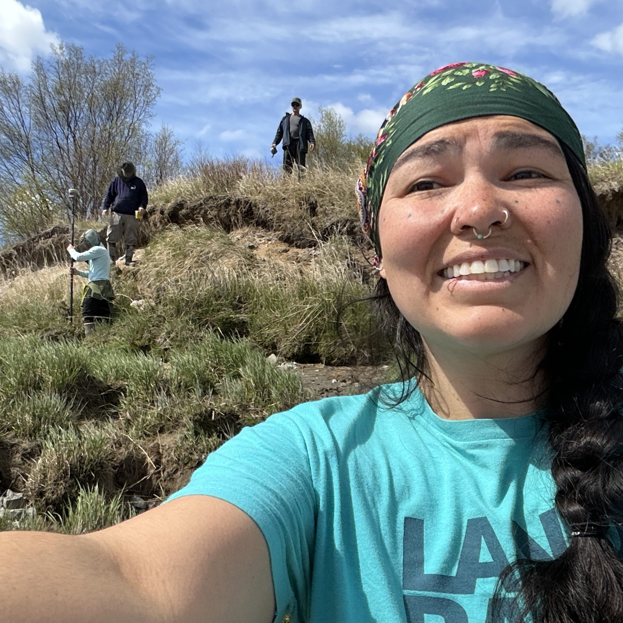 Someone is holding the camera for a selfie, behind them are three people standing at various elevations along a grassy slope. One of them is holding a Trimble instrument. The sky is cloudy. 