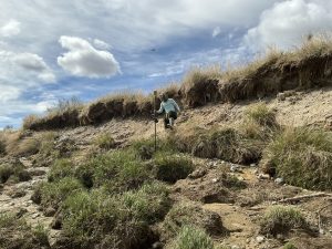 Seth taking a big step down a dirt slope while holding the Trimble instrument. The top and bottom of the slope has clumpy grass. The sky is partially cloudy.