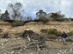 There is a coastal bluff with a fallen tree on a semi-rocky beach. Seth and Chris stand around the bluff toe on the right side of the picture. There is a grey, weathered house on the top of the bluff. There are a couple of trees around the house. The sky is partially cloudy. 