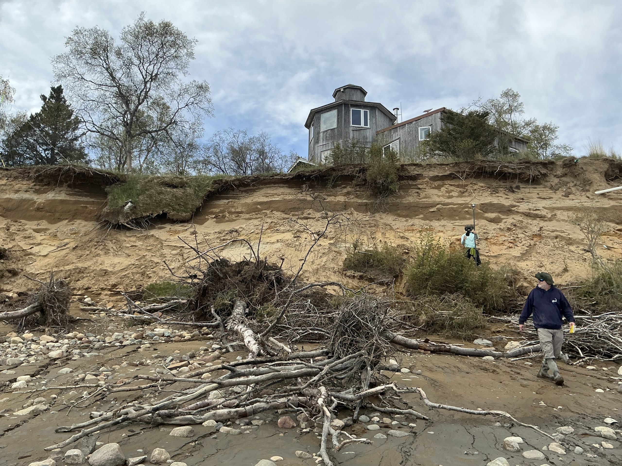 There is a coastal bluff with a fallen tree on a semi-rocky beach. Seth and Chris stand around the bluff toe on the right side of the picture. There is a grey, weathered house on the top of the bluff. There are a couple of trees around the house. The sky is partially cloudy. 