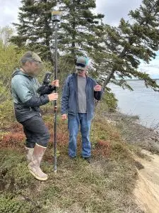 Two people stand around the top of a bluff, one of them is holding a Trimble instrument. There is tall brush and a couple of trees behind them. The sky is light grey and cloudy.