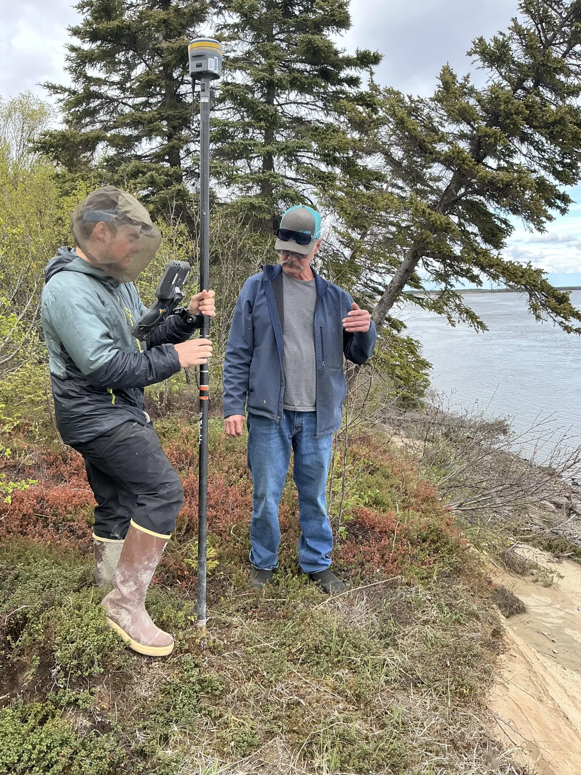 Two people stand around the top of a bluff, one of them is holding a Trimble instrument. There is tall brush and a couple of trees behind them. The sky is light grey and cloudy.