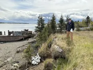 Chris stands on the top of a small grassy bluff. There are some white bouys at the bluff's toe. The beach is rocky and there is a old, rusted structure that could  be an old dock or the bottom half of a boat. There are trees in the background and the water further back appears calm and reflective. The sky is bright and cloudy.