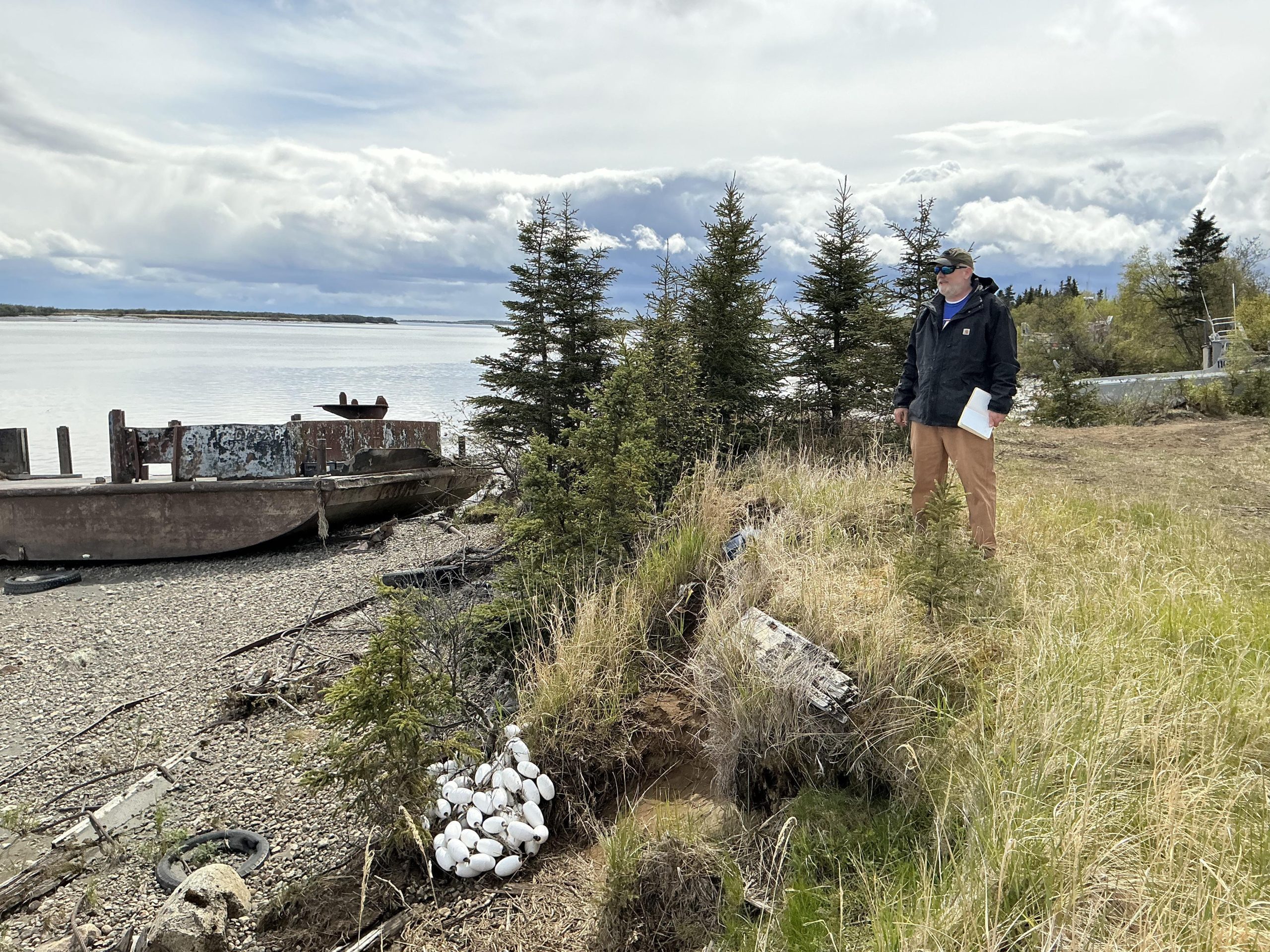 Chris stands on the top of a small grassy bluff. There are some white bouys at the bluff's toe. The beach is rocky and there is a old, rusted structure that could  be an old dock or the bottom half of a boat. There are trees in the background and the water further back appears calm and reflective. The sky is bright and cloudy.