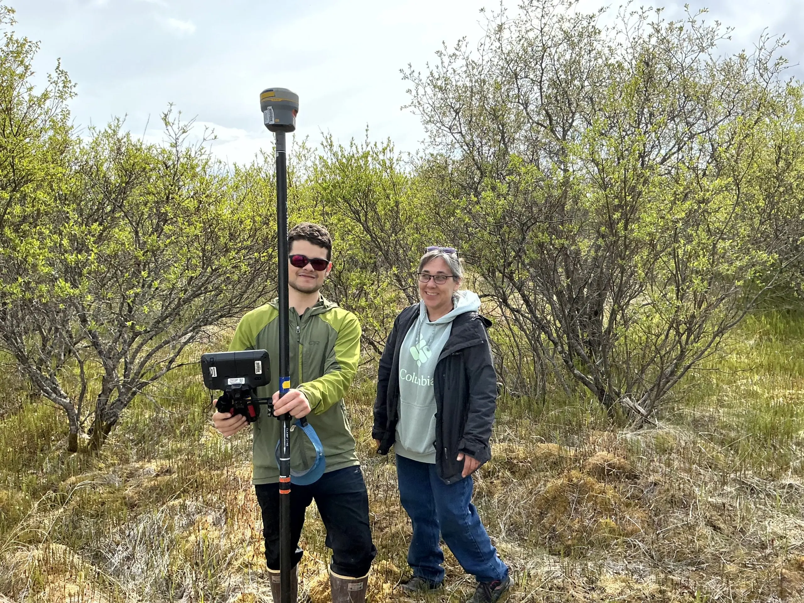 Two people stand next to each other, smiling for the photo. One of them is holding the Trimble instrument. They are standing in mossy ground with tall brush as their background. The sky is cloudy.