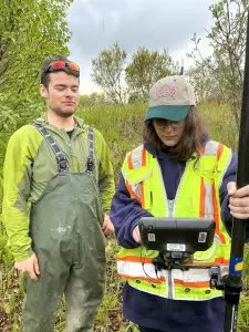 Two people stand next to each other. One of them is holding the Trimble equipment. They are standing in a grassy field with tall brush around . The sky is cloudy.