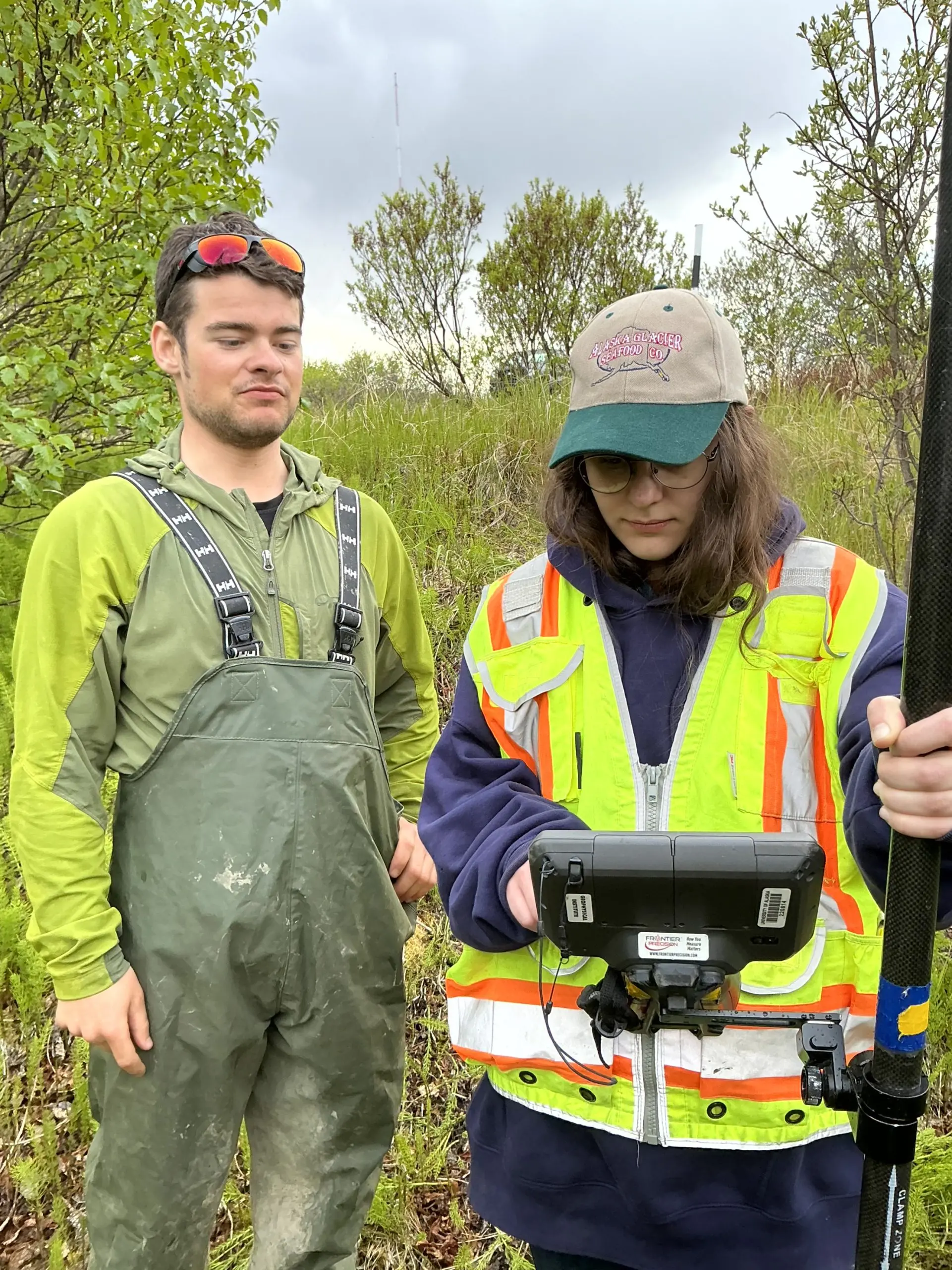 Two people stand next to each other. One of them is holding the Trimble equipment. They are standing in a grassy field with tall brush around . The sky is cloudy.