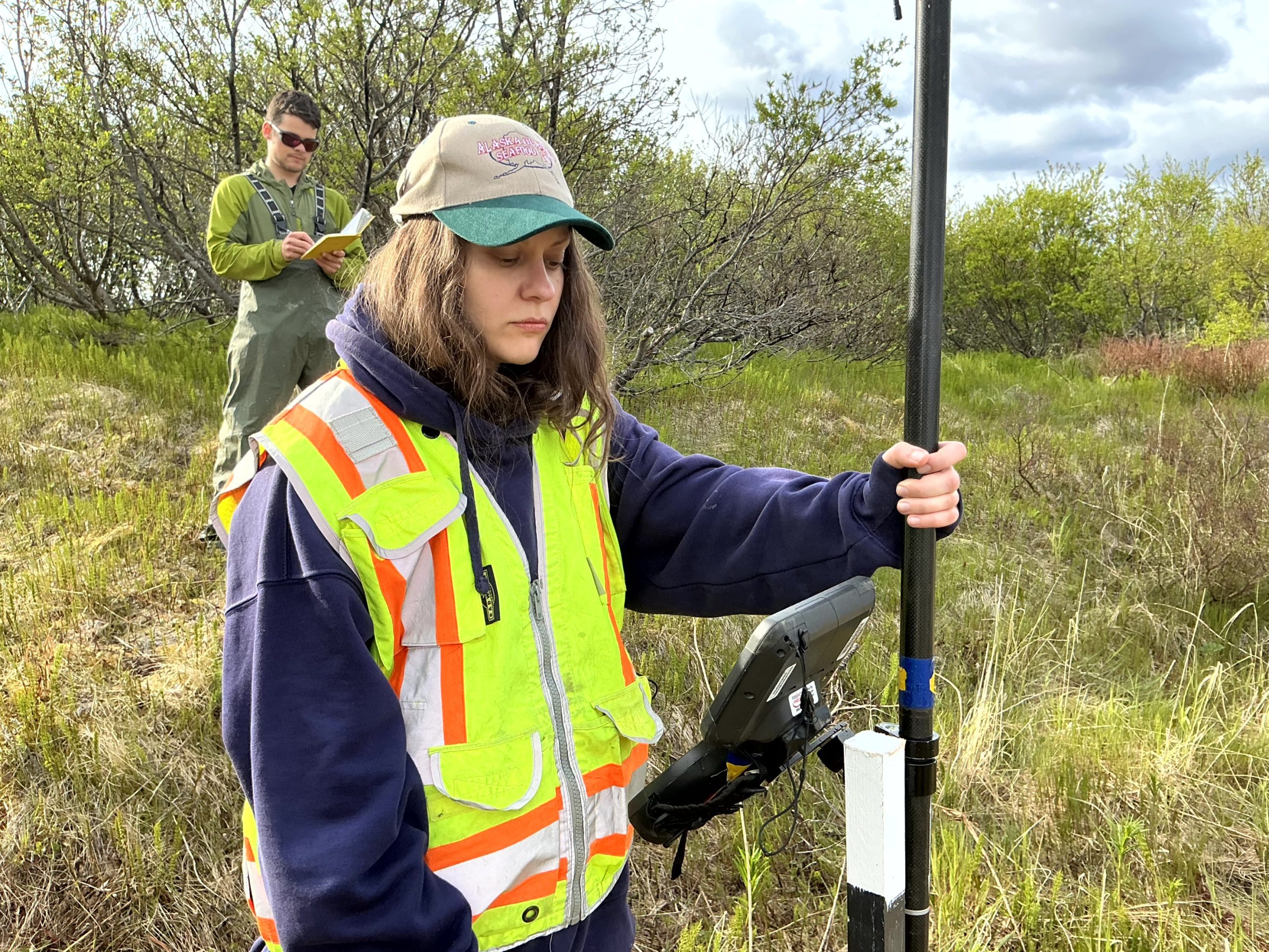 Two people stand in a grassy, hilly field. One of them is holding the Trimble instrument while the other is writing in the field notebook. There is tall brush behind them. The sky is partially cloudy.