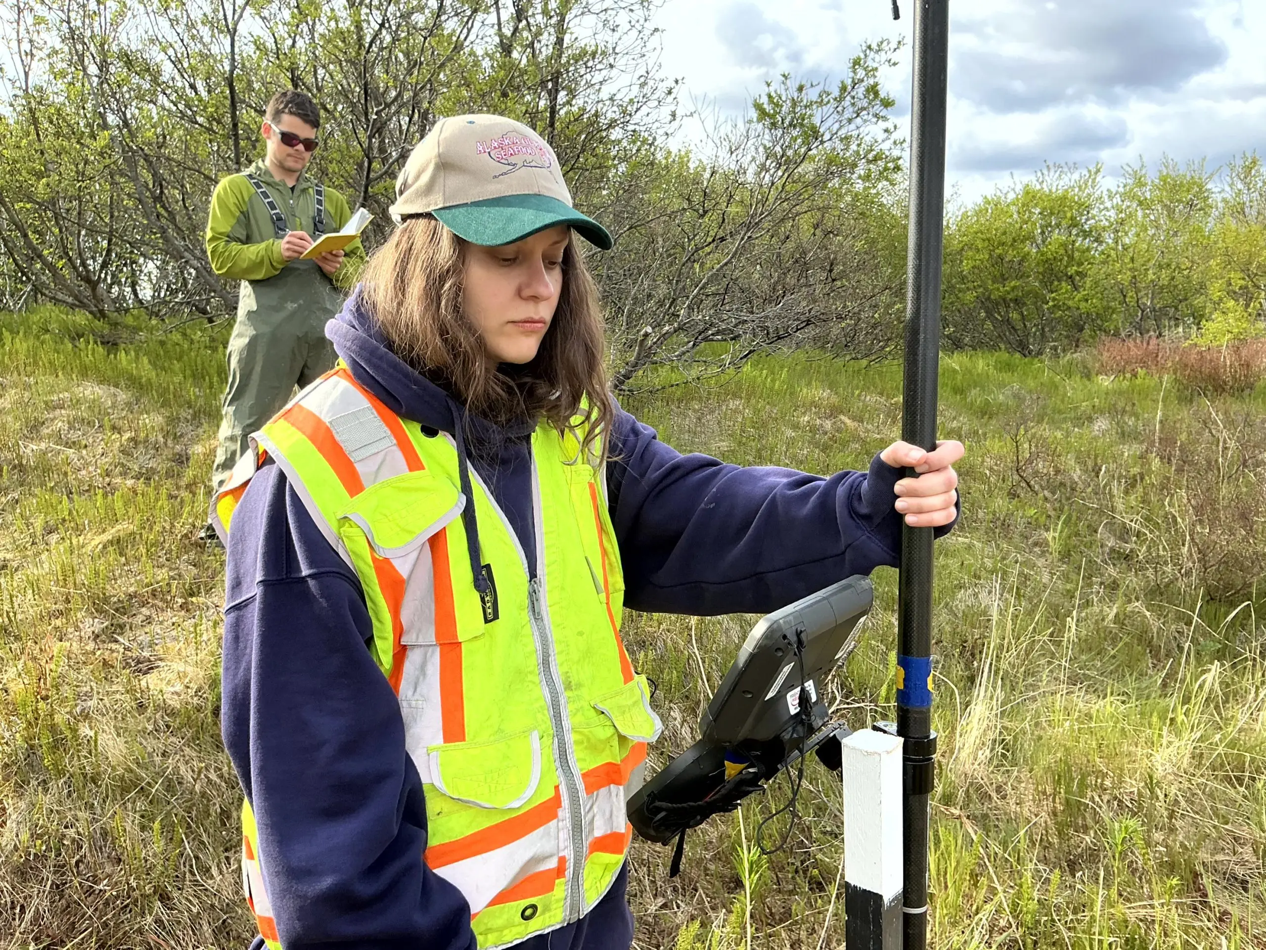Two people stand in a grassy, hilly field. One of them is holding the Trimble instrument while the other is writing in the field notebook. There is tall brush behind them. The sky is partially cloudy.