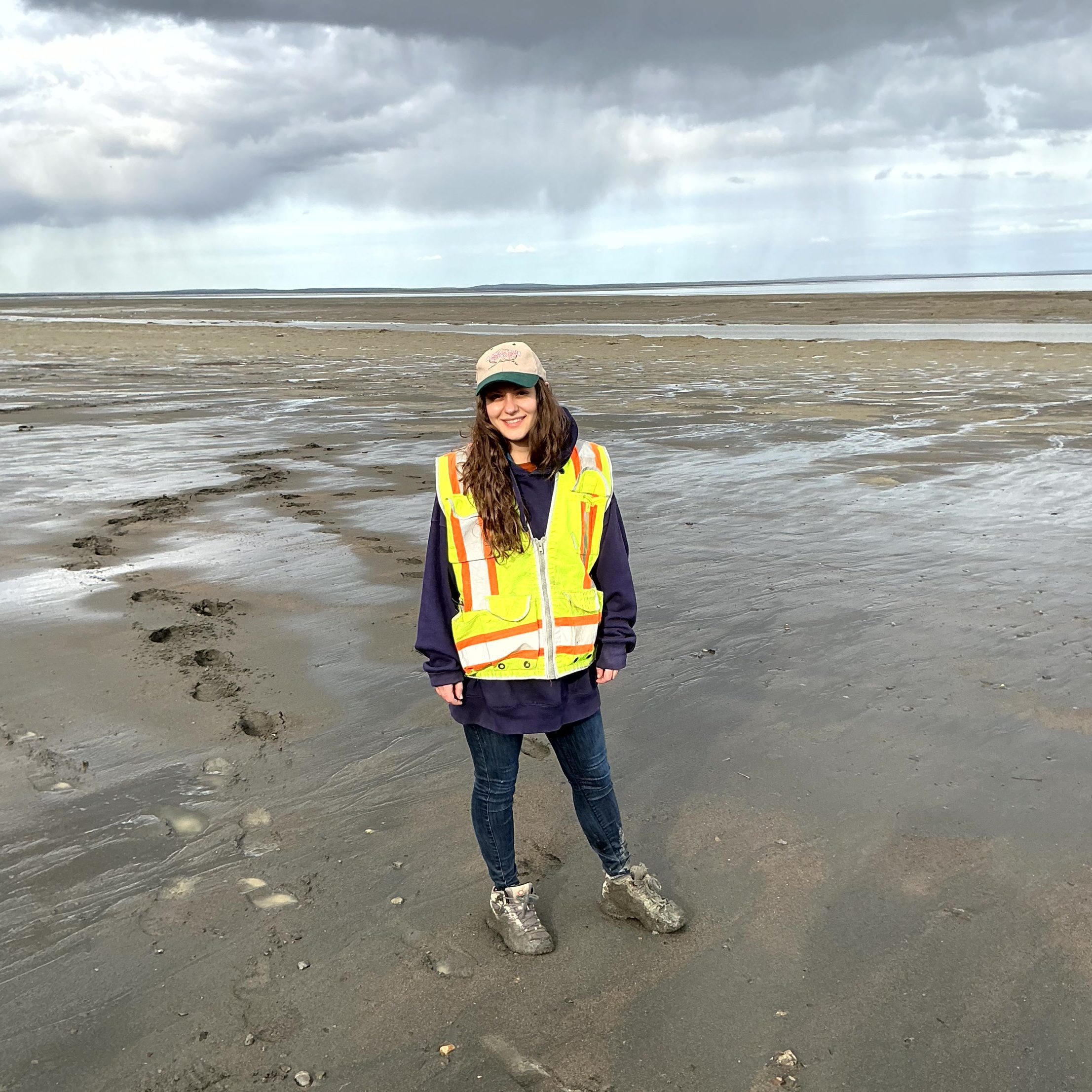 Someone stands in a large mud flat, posing for a photo. Their once white shoes are covered in mud. There are deep footprints trailing behind them heading deeper into the mud. The sky is partially cloudy and rainy.