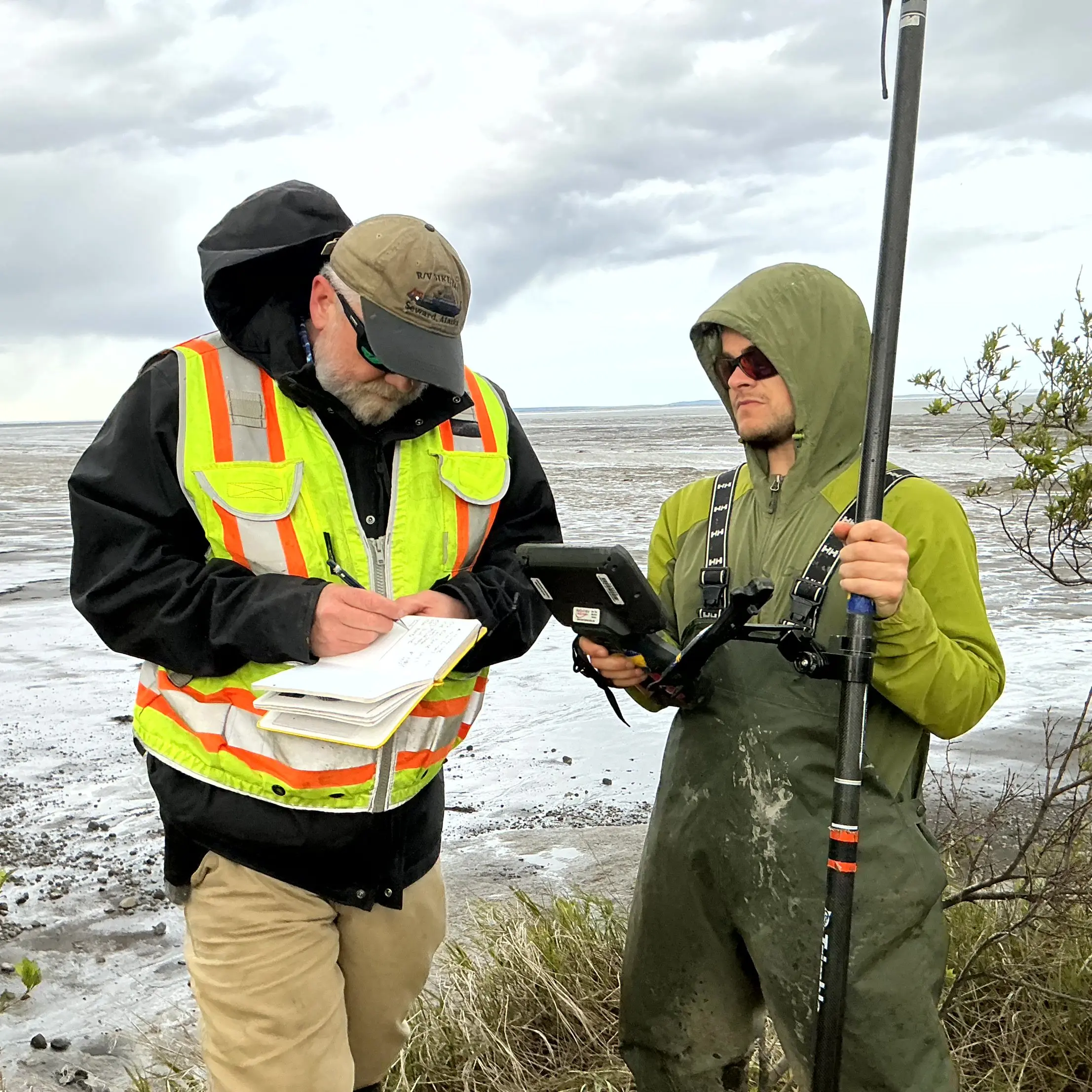 Chris and Seth stand next to each other. Chris is writing notes in the field notebook and Seth is holding the Trimble instrument. They are standing on tall grass. There is wet mud flats behind them. The sky is cloudy.