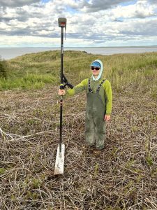 Seth stands with a Trimble instrument in a grassy field. In the background is calm waters and a partially cloudy sky. 