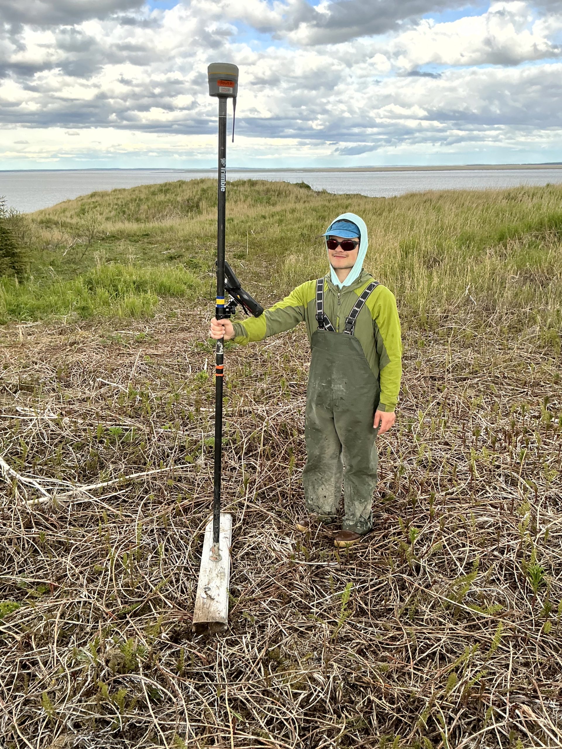Seth stands with a Trimble instrument in a grassy field. In the background is calm waters and a partially cloudy sky. 