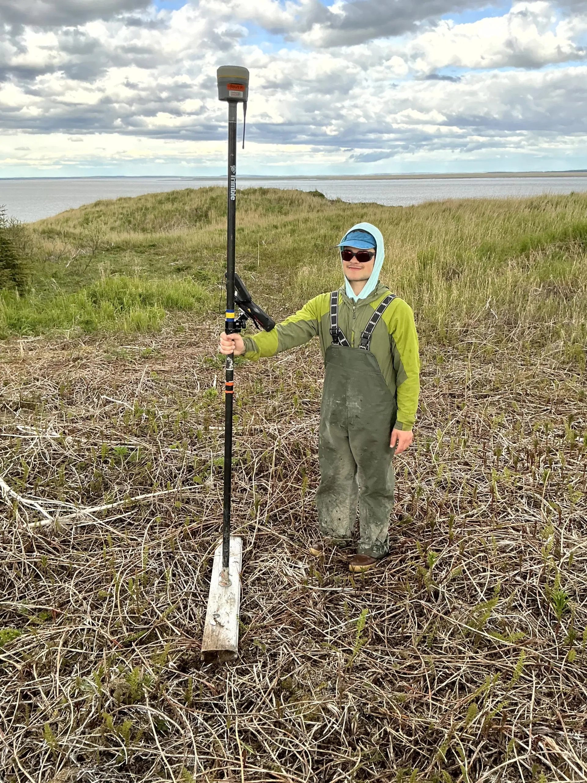 Seth stands with a Trimble instrument in a grassy field. In the background is calm waters and a partially cloudy sky. 