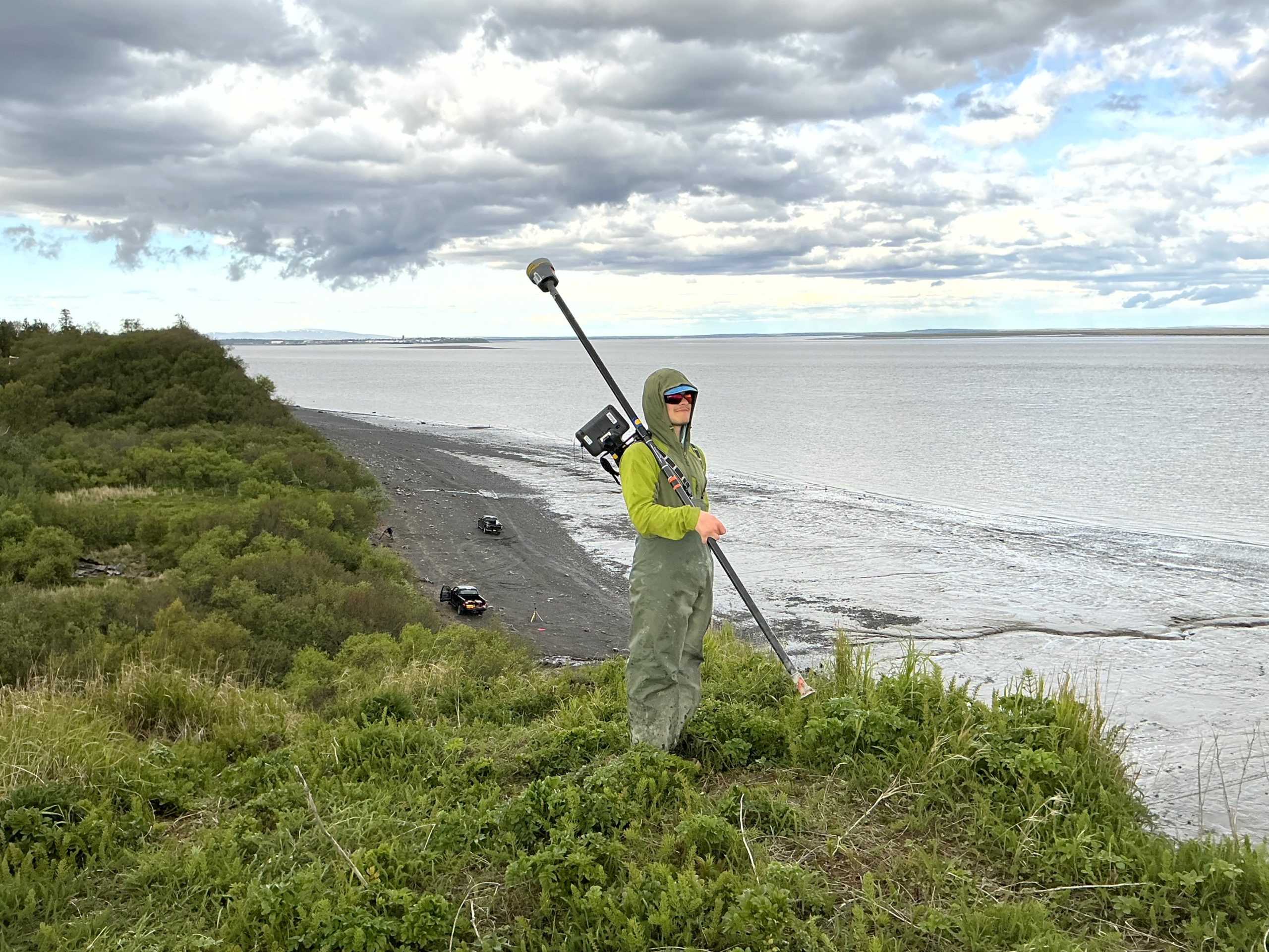 Seth stands on the top of a large bluff. He is holding a Trimble instrument. The bluff's top is green and lush. A beach can be seen at the bottom of the bluff. The water appears calm. The sky is partially cloudy. 