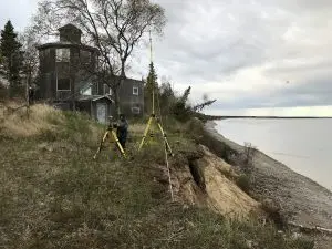 Seth is setting up the Trimble base. He is set up on top of the coastal bluff. There is an old house on the top of the bluff. The bottom of the bluff has some rocks and fallen trees. The water on the beach appears calm. The sky is cloudy.