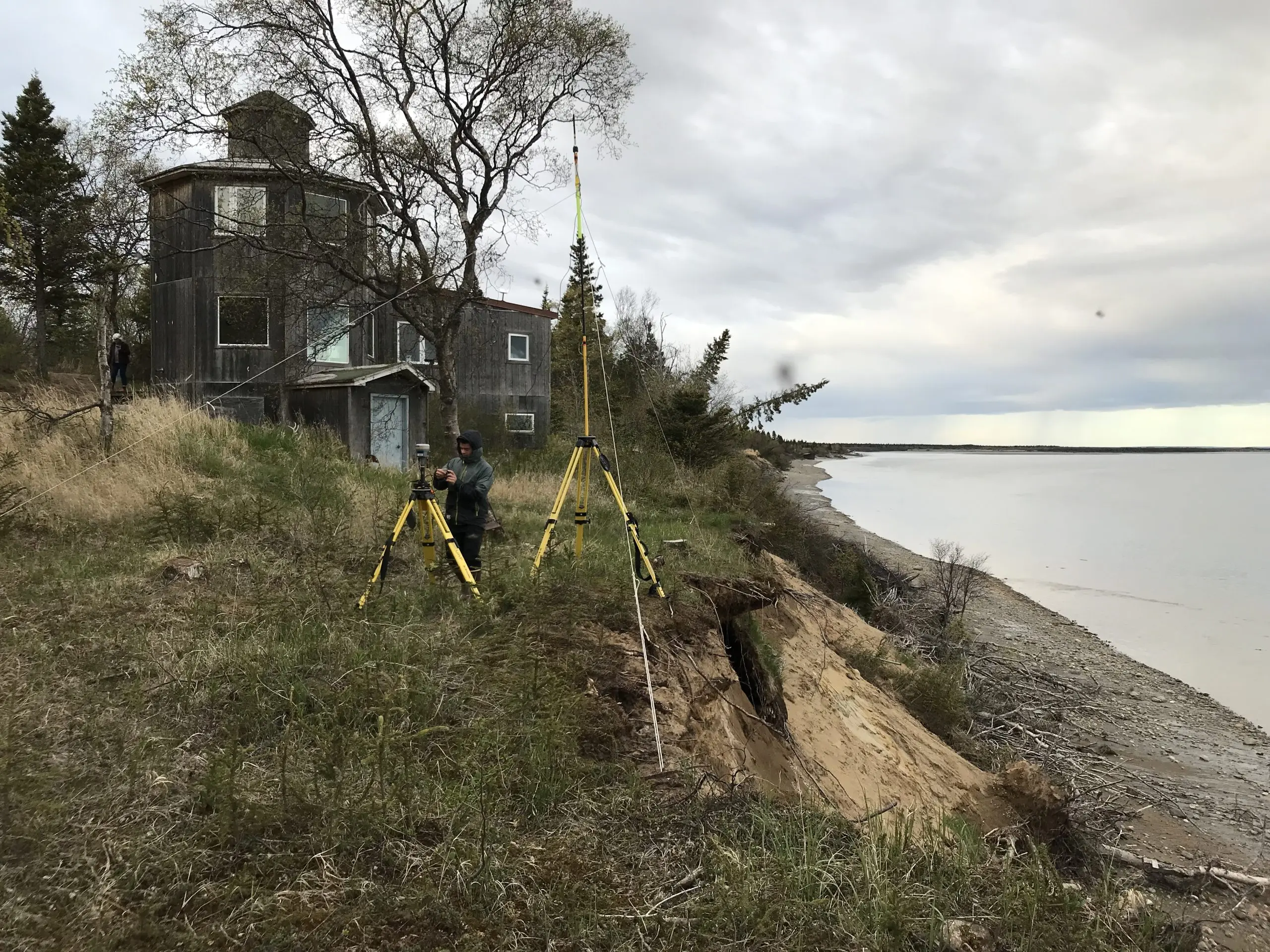 Seth is setting up the Trimble base. He is set up on top of the coastal bluff. There is an old house on the top of the bluff. The bottom of the bluff has some rocks and fallen trees. The water on the beach appears calm. The sky is cloudy.