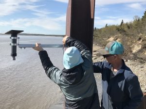 Two people are attaching a metal instrument to a large metal beam. They are above a high-tide beach. There is a small bluff covered in a mixture of brush and a few evergreen trees. The sky is partially cloudy.