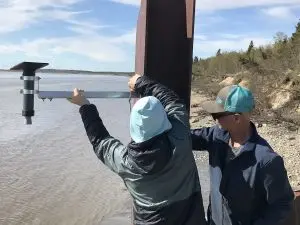 Two people are attaching a metal instrument to a large metal beam. They are above a high-tide beach. There is a small bluff covered in a mixture of brush and a few evergreen trees. The sky is partially cloudy.