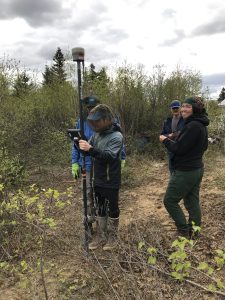 Four people stand near each other. One is holding the Trimble equipment, someone else is holding the field ipad. They are surrounded by tall brush and the sky is cloudy.
