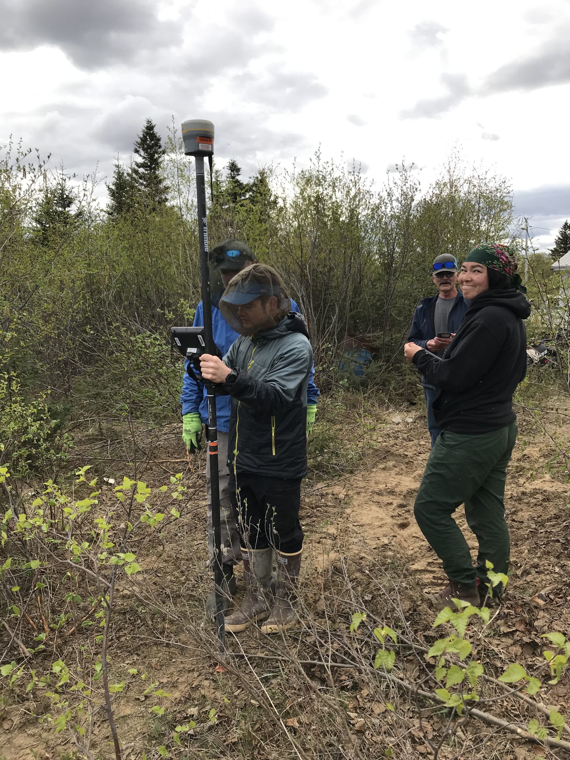 Four people stand near each other. One is holding the Trimble equipment, someone else is holding the field ipad. They are surrounded by tall brush and the sky is cloudy.