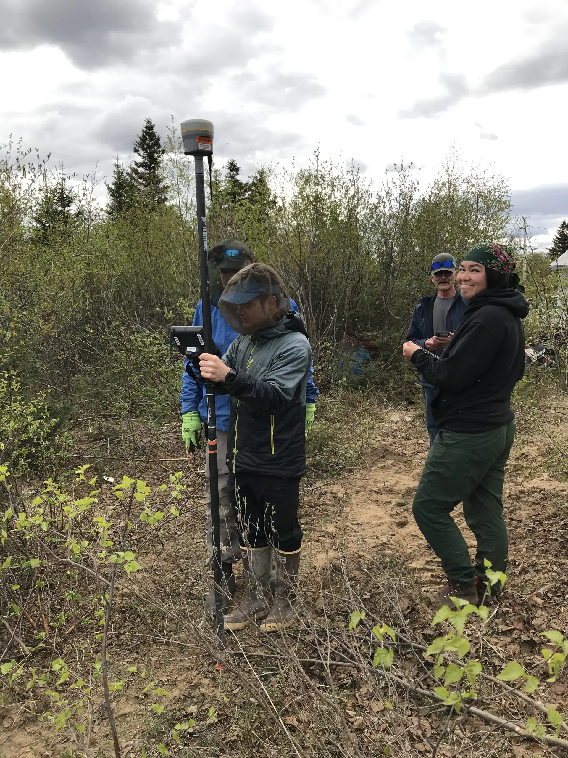 Four people stand near each other. One is holding the Trimble equipment, someone else is holding the field ipad. They are surrounded by tall brush and the sky is cloudy.