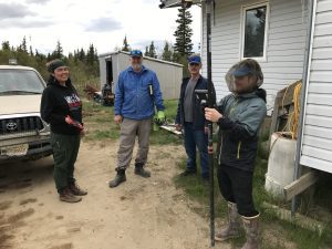 Four people stand around each other in front of a house. One of them is holding the Trimble instrument while another is holding the field ipad. There is a parked truck near one of them. There is a shed and a mixture of brush and evergreen trees in the background. The sky is cloudy. 