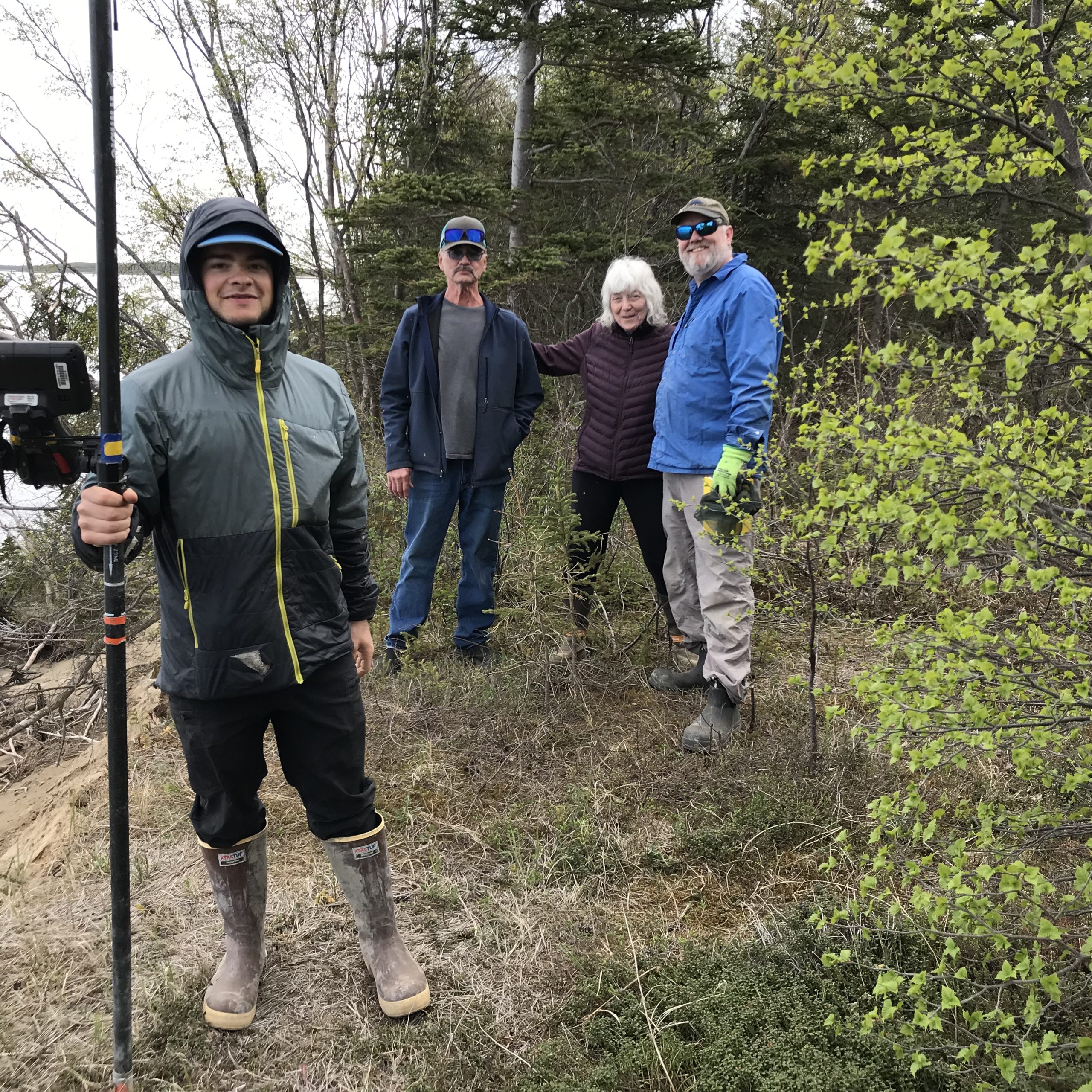 Four people stand near each other. One of them is holding the Trimble instrument. The other three are standing a little further back posing for the photo. They are standing on some grass and surrounded by trees. The sky appears cloudy. 