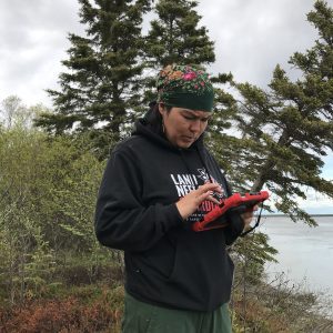Someone stands holding the field ipad. They are standing in front of some tall brush. There are some evergreen trees leaning down a bluff. Calm water can be seen in the distance. The sky is overcast.