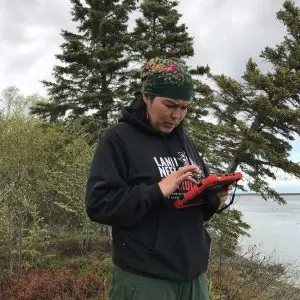 Someone stands holding the field ipad. They are standing in front of some tall brush. There are some evergreen trees leaning down a bluff. Calm water can be seen in the distance. The sky is overcast.