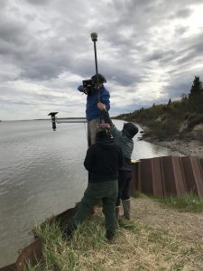 Three people stand around each other, everyone seems to be helping to elevate and balance the Trimble instrument. They are standing on a bluff that is supported by a metallic seawall. The tide in the background appears high. The coast is covered in brush and some sparse evergreen trees. The sky is overcast.
