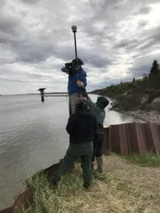 Three people stand around each other, everyone seems to be helping to elevate and balance the Trimble instrument. They are standing on a bluff that is supported by a metallic seawall. The tide in the background appears high. The coast is covered in brush and some sparse evergreen trees. The sky is overcast.