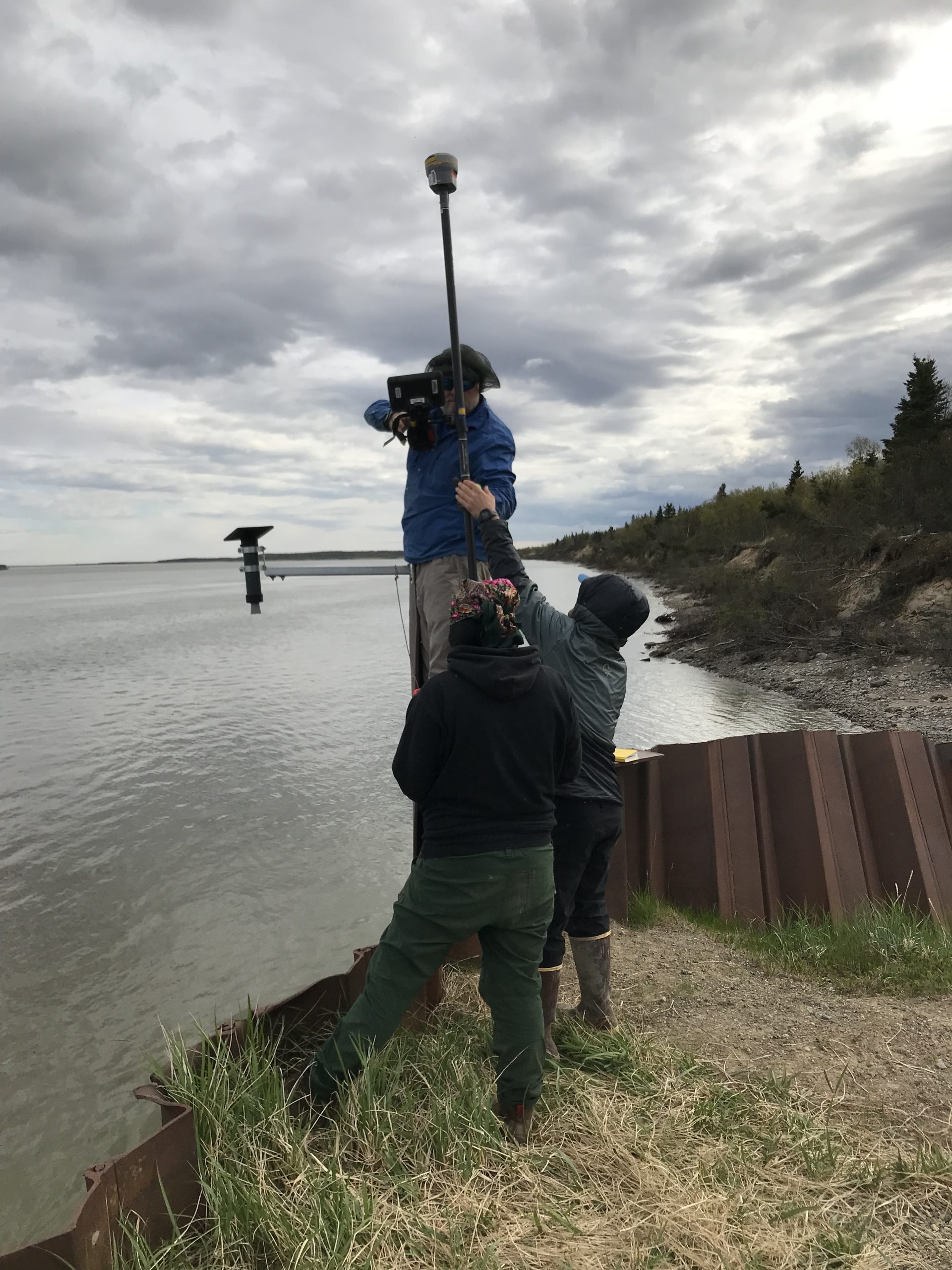 Three people stand around each other, everyone seems to be helping to elevate and balance the Trimble instrument. They are standing on a bluff that is supported by a metallic seawall. The tide in the background appears high. The coast is covered in brush and some sparse evergreen trees. The sky is overcast.