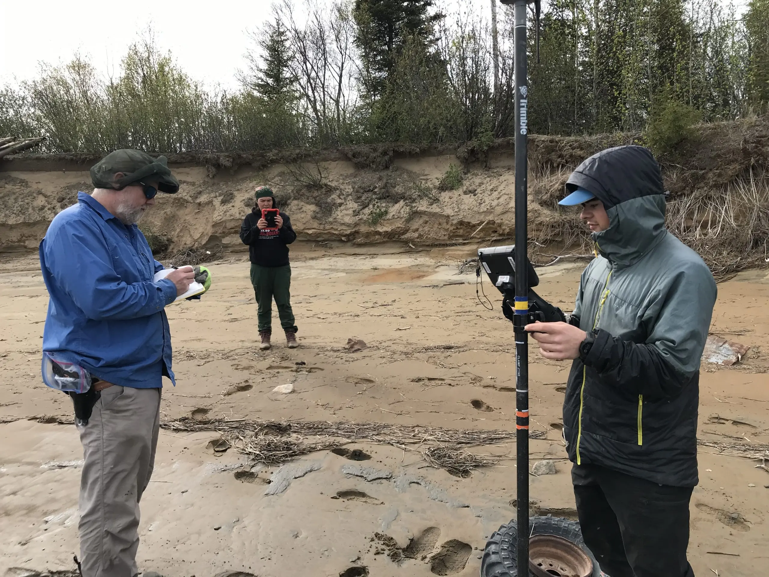 Three people stand around a muddy beach. One is holding a Trimble instrument, another is taking field notes, and the third is taking a photo with the ipad. There is a bluff behind them. On top of the bluff are some trees and brush. The sky is overcast.