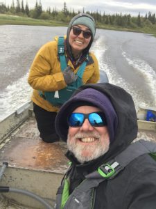 Two people are sitting in a moving boat. They are looking toward the camera and smiling. There are motion wakes in the water behind the boat. The shore is covered in green brush and trees. The sky is overcast.