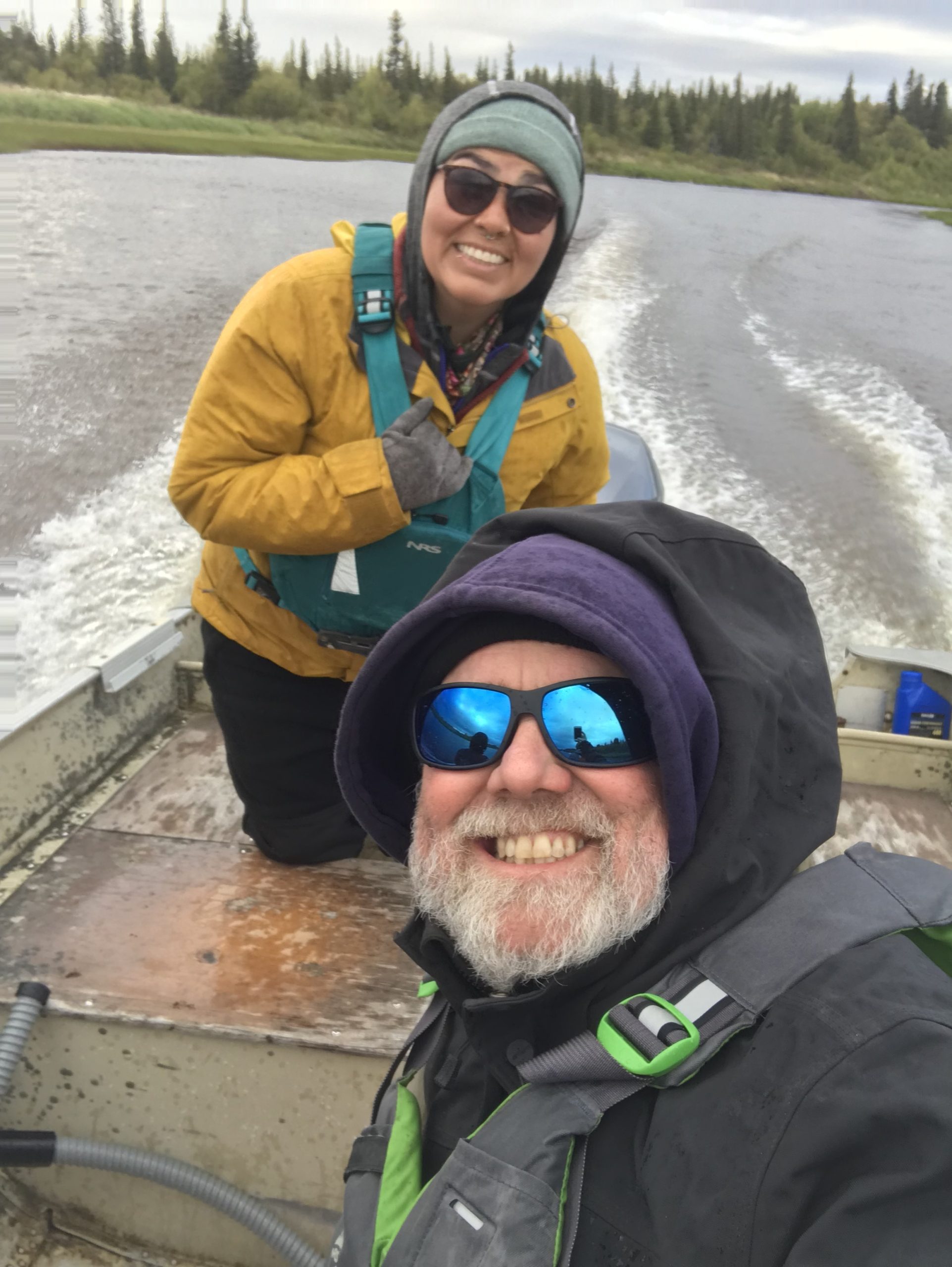 Two people are sitting in a moving boat. They are looking toward the camera and smiling. There are motion wakes in the water behind the boat. The shore is covered in green brush and trees. The sky is overcast.