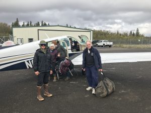 Four people are standing around a small airplane and an extra person is sitting in the plane's cockpit. One person is loading luggage into the plane. Three other people are looking at the camera and smiling. They are standing on asphalt. There is a small building in the background and chain link fencing. Beyond the fencing are trees and the sky is overcast.