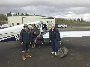 Four people are standing around a small airplane and an extra person is sitting in the plane's cockpit. One person is loading luggage into the plane. Three other people are looking at the camera and smiling. They are standing on asphalt. There is a small building in the background and chain link fencing. Beyond the fencing are trees and the sky is overcast.