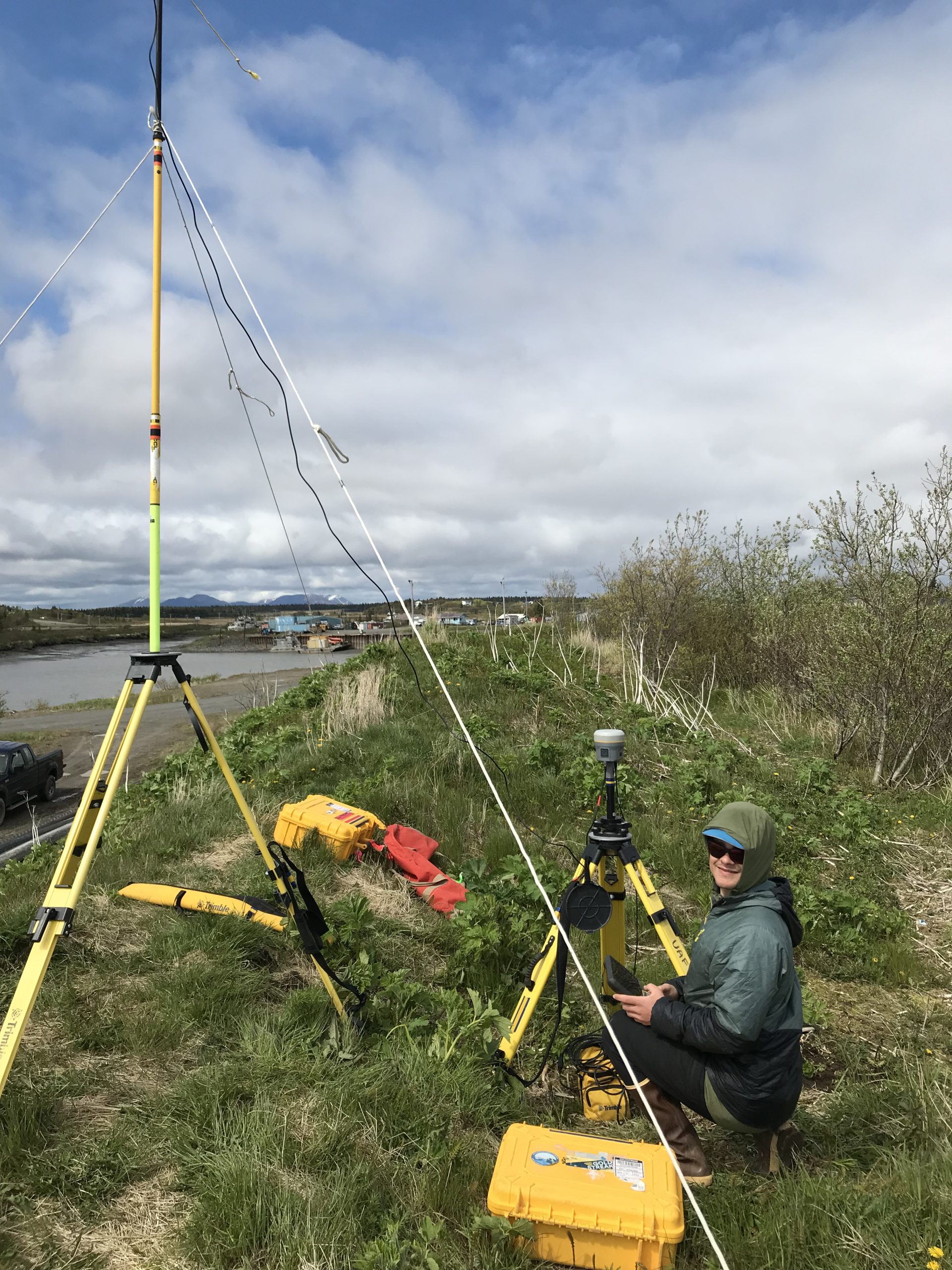 Seth Classen sits next to the Trimble base equipment. They are on top of a grassy hill. The background is a harbor. The sky is partially cloudy. 