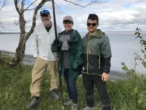 Three people stand next to each other smiling at the camera. One of them is holding the Trimble instrument. They are standing on top of a grassy bluff. There are calm ocean waters in the background and a partially cloudy sky. 