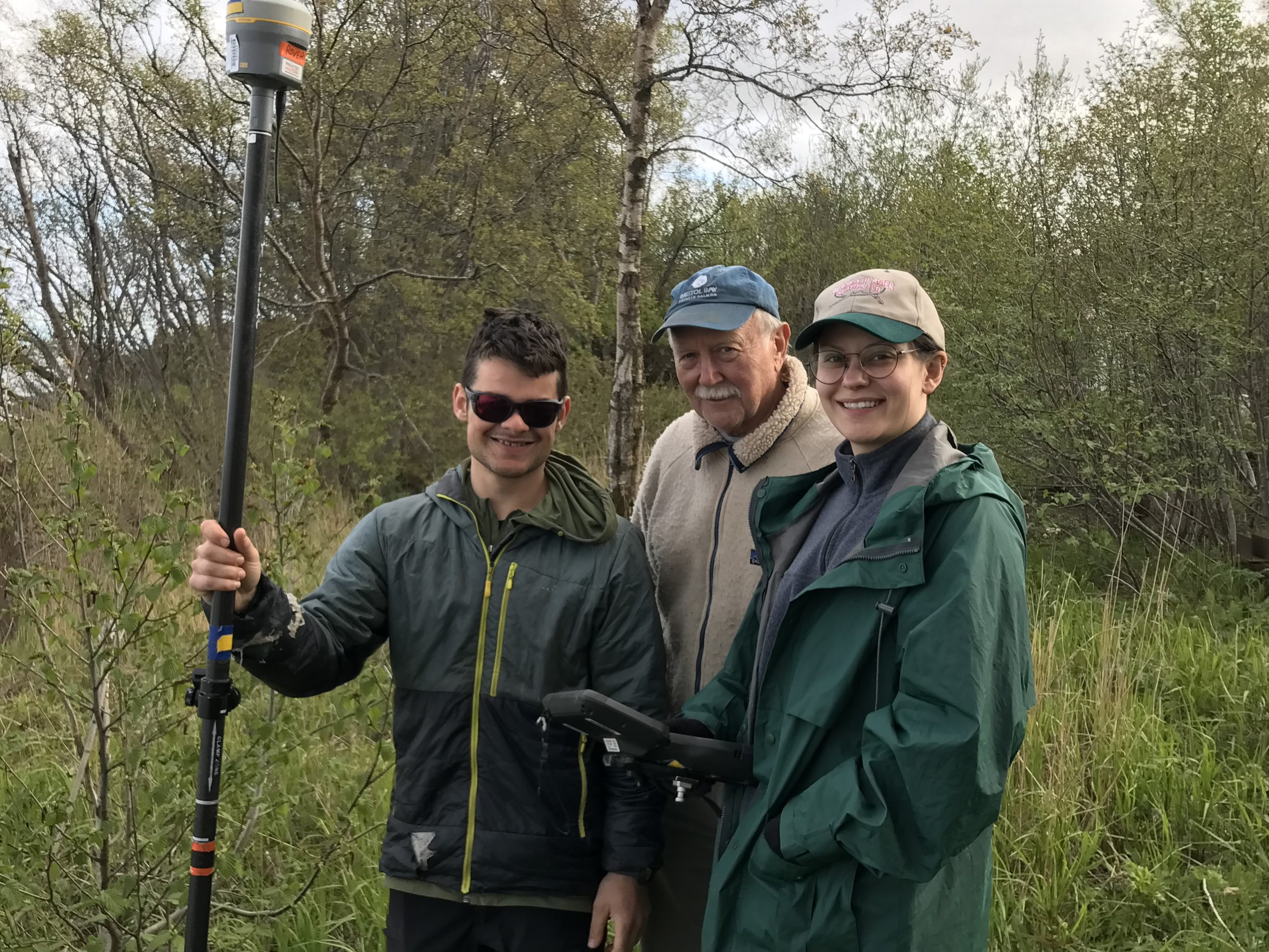Three people standing next to each other smiling at the camera. One of them is holding the Trimble stick while the other is holding the Trimble controller. They are standing in a grassy area surrounded by brush. The sky appears mostly overcast.