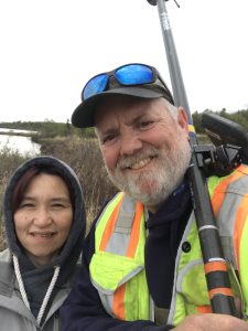 Two people standing next to each other for a selfie. One of them is holding the Trimble instrument. They are standing in a vegetated area near the coast. The sky is overcast. 