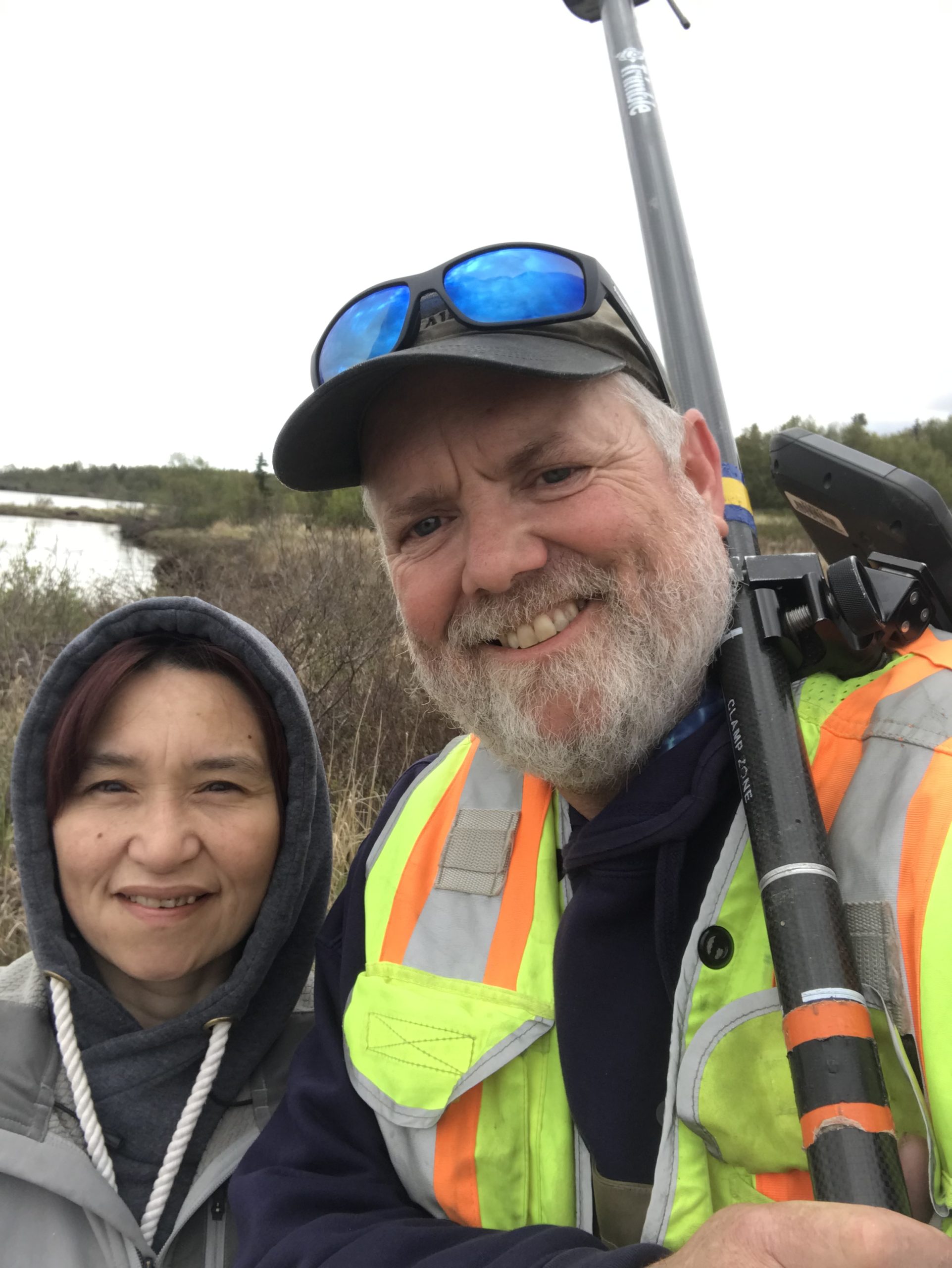 Two people standing next to each other for a selfie. One of them is holding the Trimble instrument. They are standing in a vegetated area near the coast. The sky is overcast. 