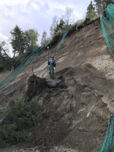 Seth Classen stands in the middle of a tall bluff. Green netting is draped around various locations around the bluff. There is grass and a couple of trees around the top of the bluff. The sky is cloudy. 