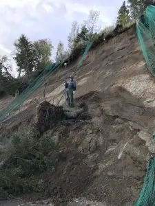 Seth Classen stands in the middle of a tall bluff. Green netting is draped around various locations around the bluff. There is grass and a couple of trees around the top of the bluff. The sky is cloudy. 