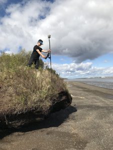 Seth Classen stands on top of a small bluff holding a Trimble instrument. The tide on the beach below is low. The sky is cloudy. 